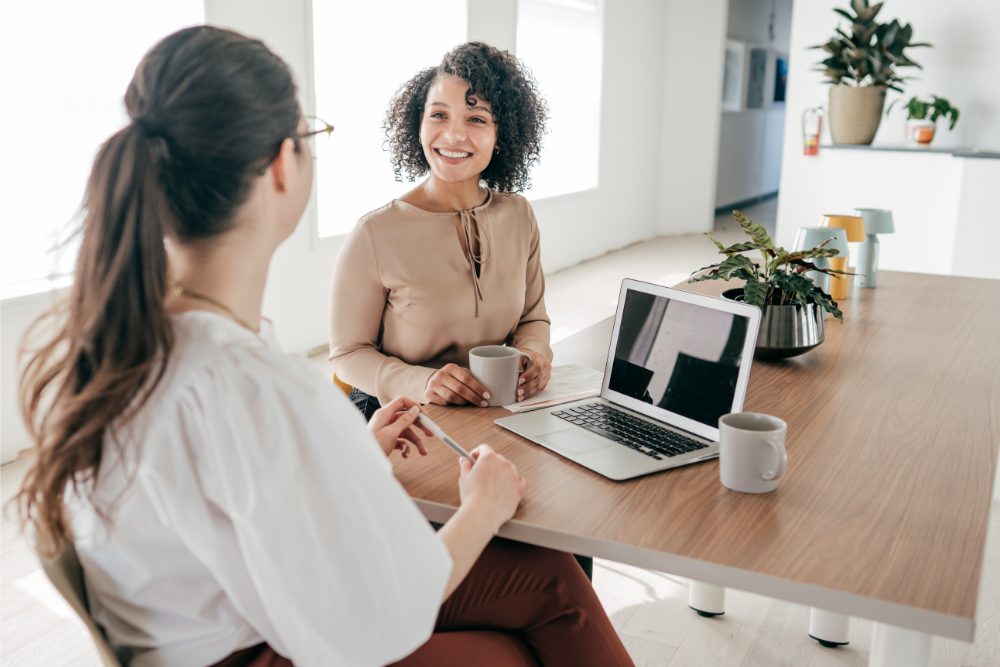 HR can help employees grow while WFH by giving them plenty of praise and recognition. Photo of two women sitting and smiling.